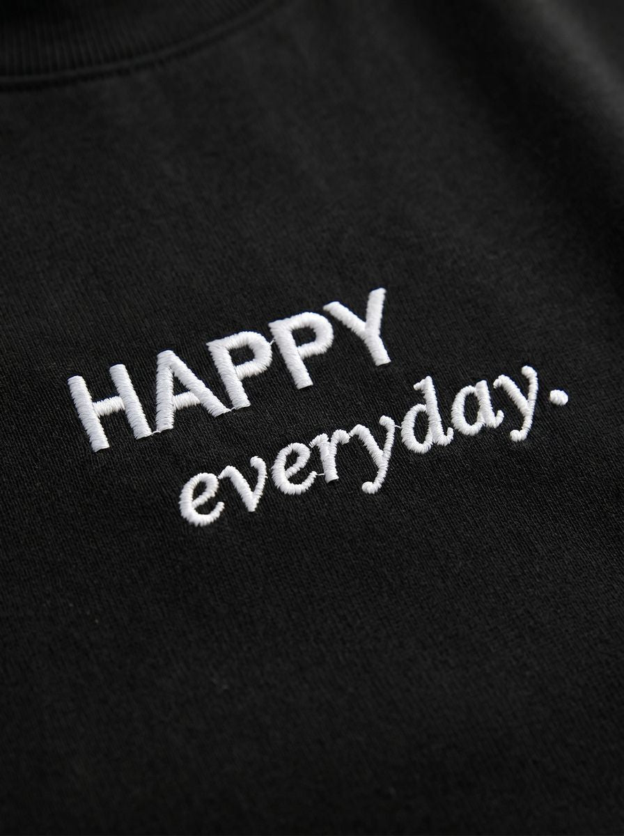 Kokybiško balto siuvinėjimo „HAPPY everyday.“ detalė ant juodų moteriškų marškinėlių.